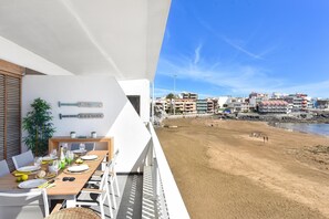 Un balcón con una mesa de comedor lista para cuatro personas, con vista a una playa de arena y al paisaje urbano.
