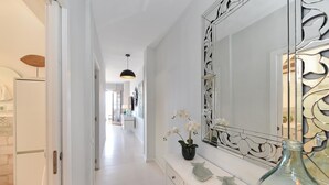 Een hal met een witte koelkast, een wit kastje met lades, een grote spiegel met decoratieve details, een vaas met witte bloemen, en een glazen fles.