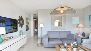 Een moderne woonkamer met een grijze bank, een bijzettafeltje met decoratieve objecten, een televisie op een witte kast, en een grote spiegel boven de bank.