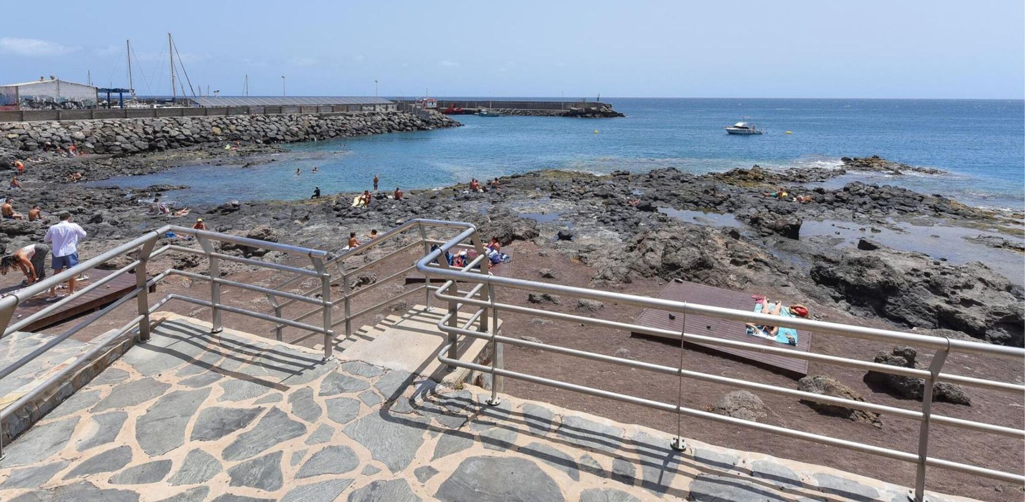 Une zone côtière avec un escalier en pierre menant à une plage, une côte rocheuse et une jetée en arrière-plan.