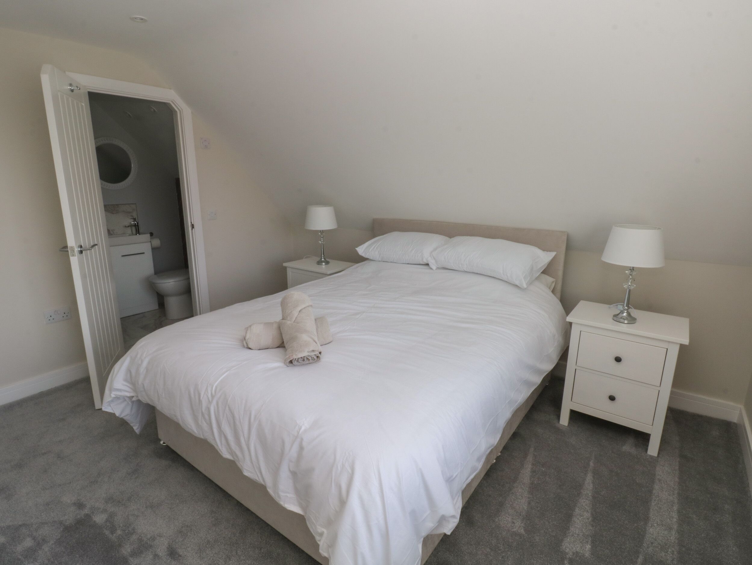 A bedroom with a bed, two bedside tables with lamps, a small bathroom with a toilet and sink, and a door leading to another room.
