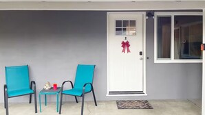 Exterior de una casa con una puerta blanca marcada con el número 2, dos sillas turquesa, una mesita con una taza roja y un felpudo.
