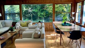 A spacious living room with a wooden floor, a sofa, a dining table with chairs, and large windows offering a view of trees. - Spacious 3 BDR 3 BA 2 Acre property one minute from Lake Michigan-Dog Friendly! (Coloma)