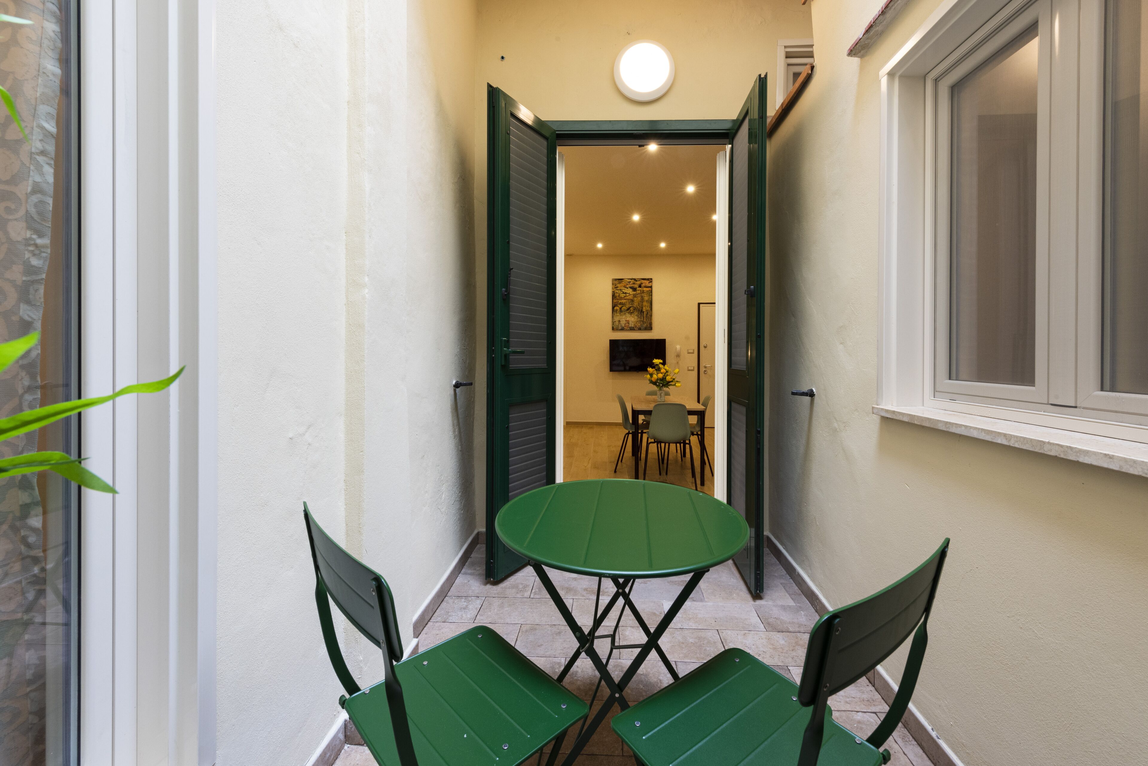 A narrow corridor with a green table and chairs, leading to a room with a TV and a painting.