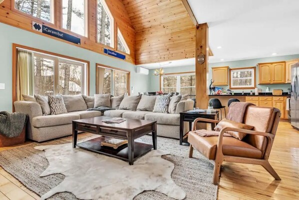 A spacious living room with a large sectional sofa, a leather armchair, and a coffee table. There are multiple windows providing natural light and a view of the surrounding forest.