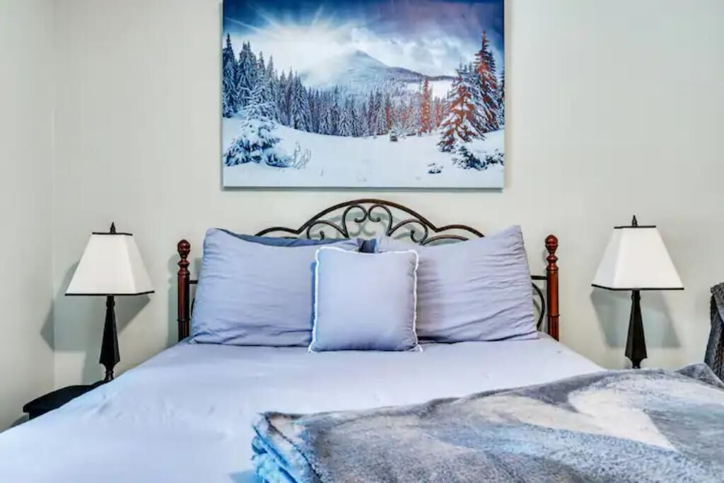 A neatly made bed with a mountain landscape painting above it, two bedside lamps, and a headboard with a decorative design.