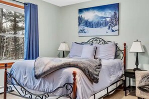 A bedroom with a bed, two bedside tables, a window with curtains, a painting on the wall, and a chair.