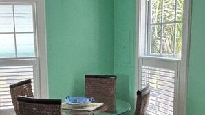 A dining area with a round glass table surrounded by wicker chairs, set against a green wall with windows. - Large 1 bedroom condo in the historic area of Key West! (Key West)