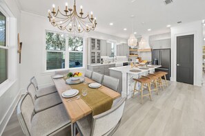 A modern kitchen with a central island, a dining area with a round table, and a large window providing a view of the outdoors. - "White Heron" - Perfectly Located on the West End of Panama City Beach w/ Private Pool + easy 5 minute walk to the Beach! (Panama City Beach)