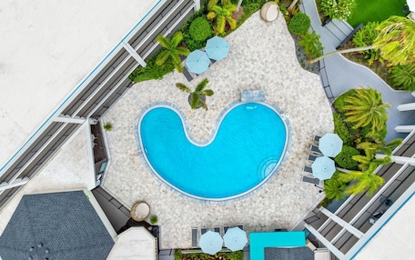 An aerial view of a hotel with a heart-shaped pool surrounded by lounge chairs and umbrellas, with palm trees and a landscaped garden.