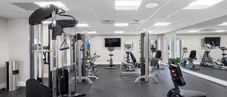 A gym with various exercise machines, a TV on the wall, and a mirror reflecting the equipment.