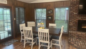 A dining room with a wooden table and white chairs, large windows, and a brick fireplace.