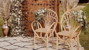 Uma porta de madeira rústica com detalhes metálicos decorativos, ladeada por duas cadeiras de vime e rodeada por flores secas e folhagem.