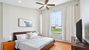 Une chambre à coucher avec un lit, un ventilateur de plafond, une télévision, une table de chevet et une fenêtre avec des rideaux.