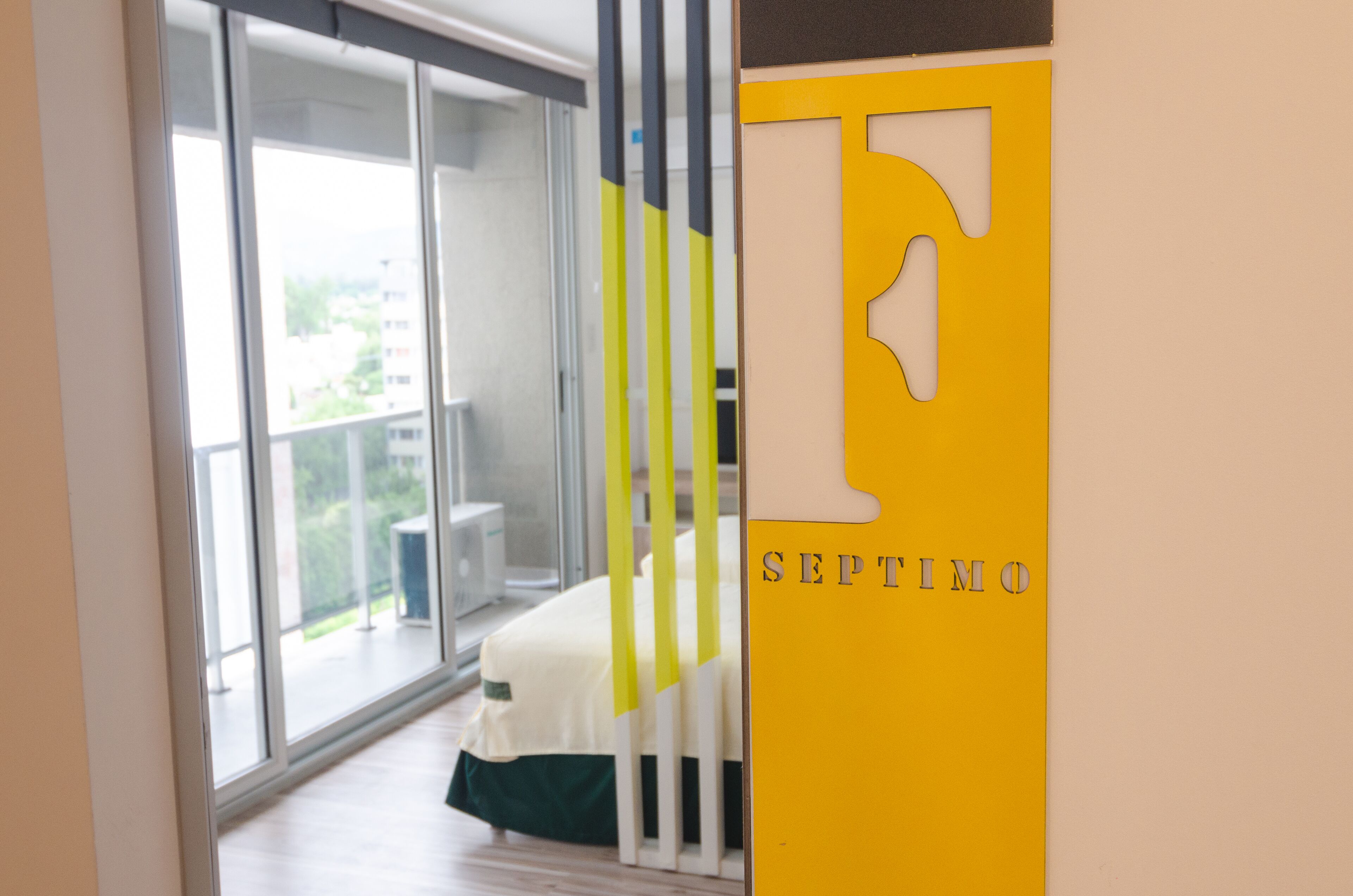A modern hotel room with a large sliding glass door leading to a balcony, a bed with a green and white bedspread, and a yellow wall with a large 'F' and the word 'SEPTIMO'.