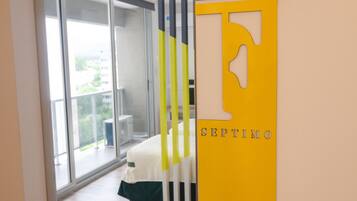 A modern hotel room with a large sliding glass door leading to a balcony, a bed with a green and white bedspread, and a yellow wall with a large 'F' and the word 'SEPTIMO'.
