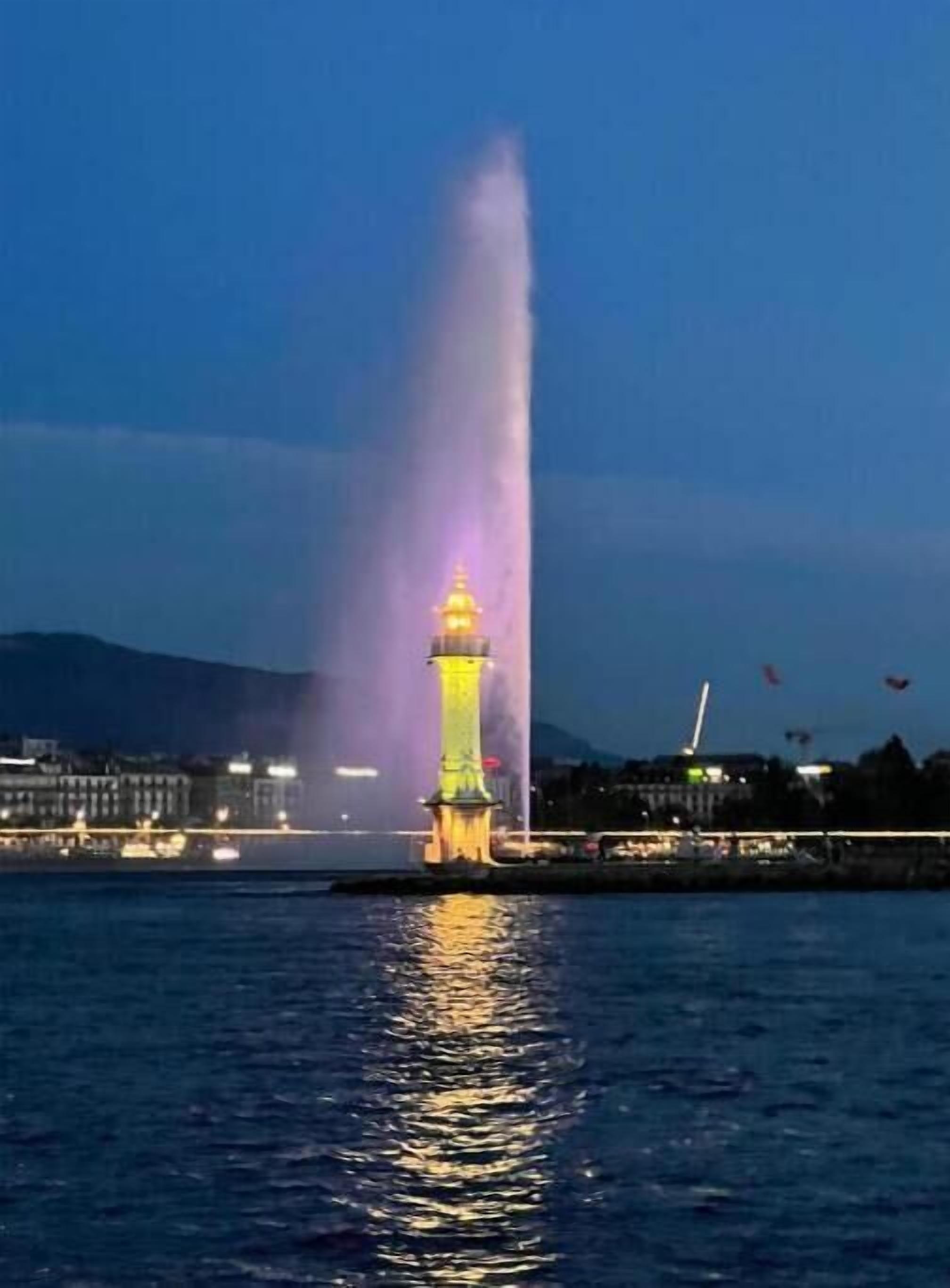 A lighthouse with a water jet in the foreground, a cityscape in the background, and a clear night sky.