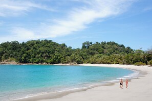Ein Strand mit klarem türkisblauem Wasser, sandigem Ufer und üppiger grüner Vegetation im Hintergrund.