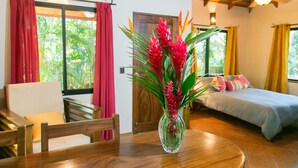A room with a bed, a dining table with chairs, a vase with flowers, and a window with curtains. - Piece of Paradise in Manuel Antonio 7 Minutes to the Beach! Casa Colibri (Quepos)