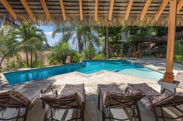 A swimming pool with lounge chairs under a thatched roof, surrounded by tropical vegetation. - Piece of Paradise in Manuel Antonio 7 Minutes to the Beach! Casa Colibri (Quepos)