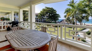 Een balkon met een houten tafel en stoelen, met uitzicht op een strand met palmbomen en een zeezicht.