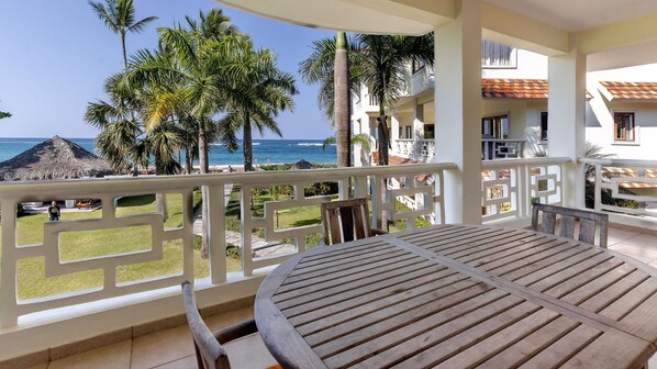 Een balkon met een houten tafel en stoelen, met uitzicht op een strand met palmbomen en een zeezicht.