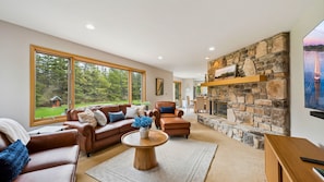 A spacious living room with a stone fireplace, leather sofas, and large windows offering a view of the outdoors. - Charming Sister Bay Home | Backyard Oasis & HotTub (Sister Bay)