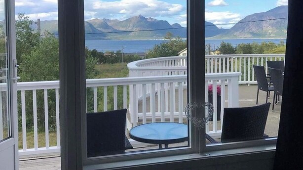 Une pièce dotée d’une grande fenêtre donnant sur une terrasse aménagée avec des sièges et offrant une vue panoramique sur les montagnes et un lac.