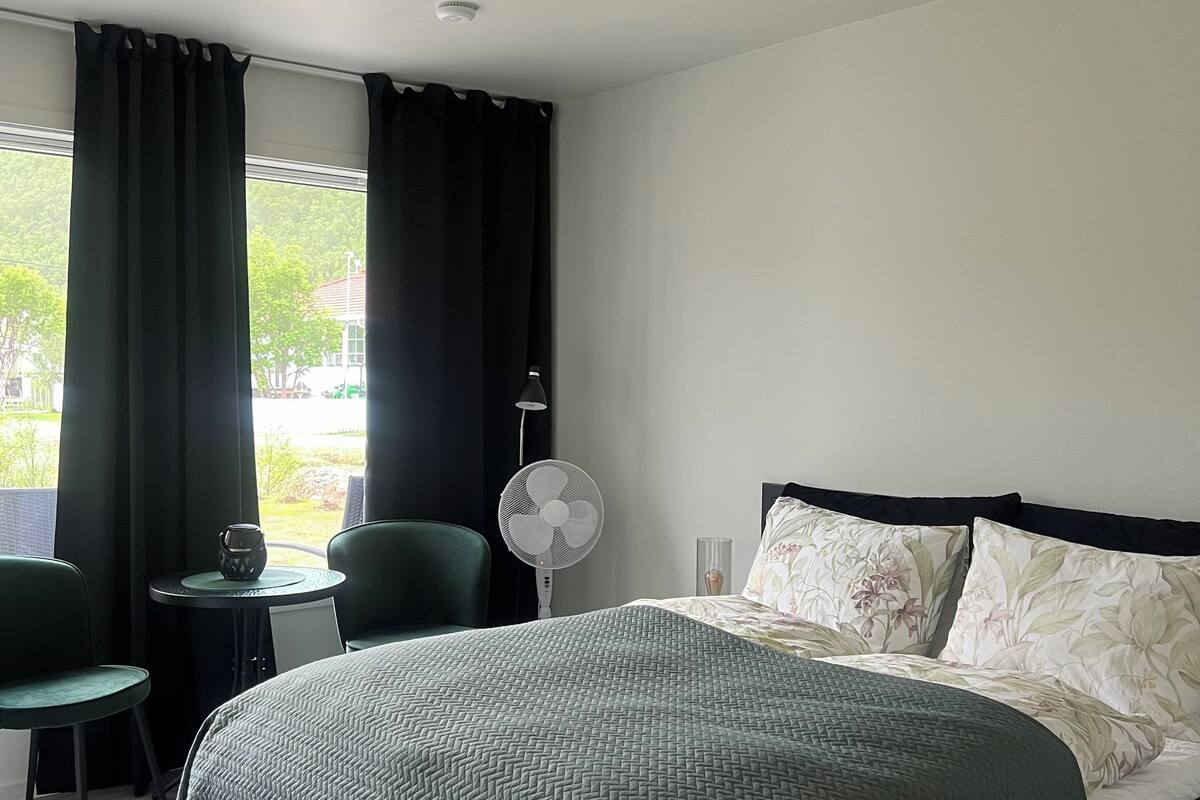 Une chambre d’hôtel avec un lit, une petite table sur laquelle se trouve un ventilateur, une chaise et une vue sur l’extérieur.