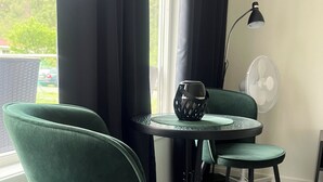 Une pièce comprenant deux fauteuils verts, une petite table ronde, un lampadaire et un ventilateur sur pied.