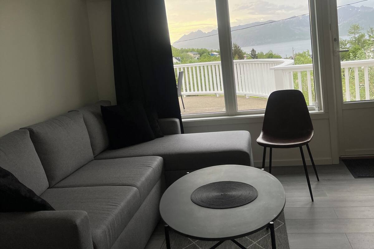 Un salon moderne avec un canapé gris, une petite table ronde et une vue sur les montagnes et un lac, visible à travers de grandes fenêtres.