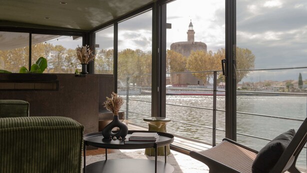 A modern room with a large window overlooking a river, a view of a historic tower, and a seating area with a table and chairs.