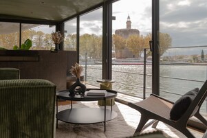Una habitación moderna con un ventanal grande que da al río, vista de una torre histórica, y un área de descanso con mesa y sillas.