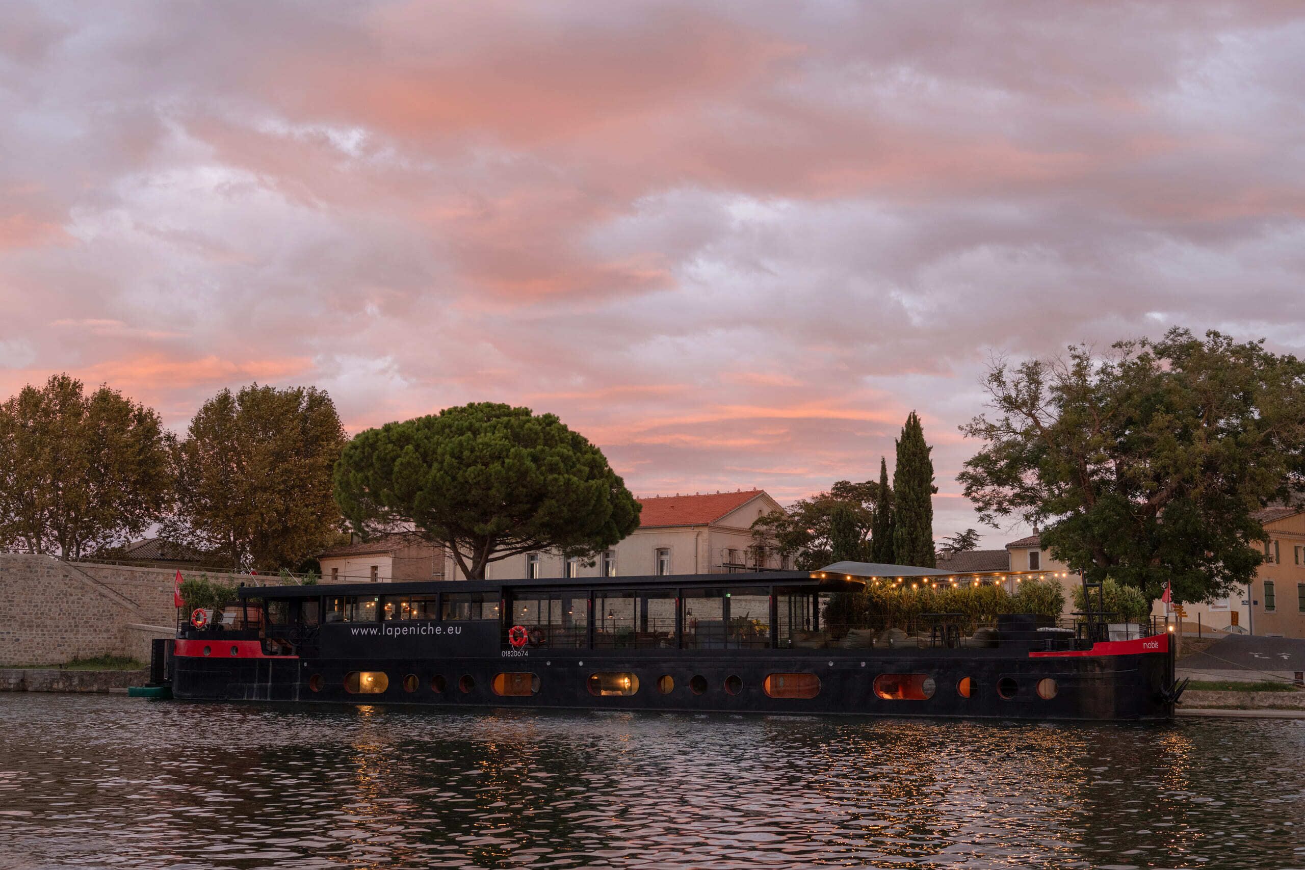 A boat with a website URL and a lifebuoy, docked at a waterfront with trees and buildings in the background.