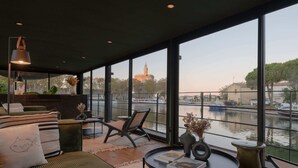 A modern interior with large windows overlooking a canal, featuring a sofa, chairs, a small table, and a bookshelf.