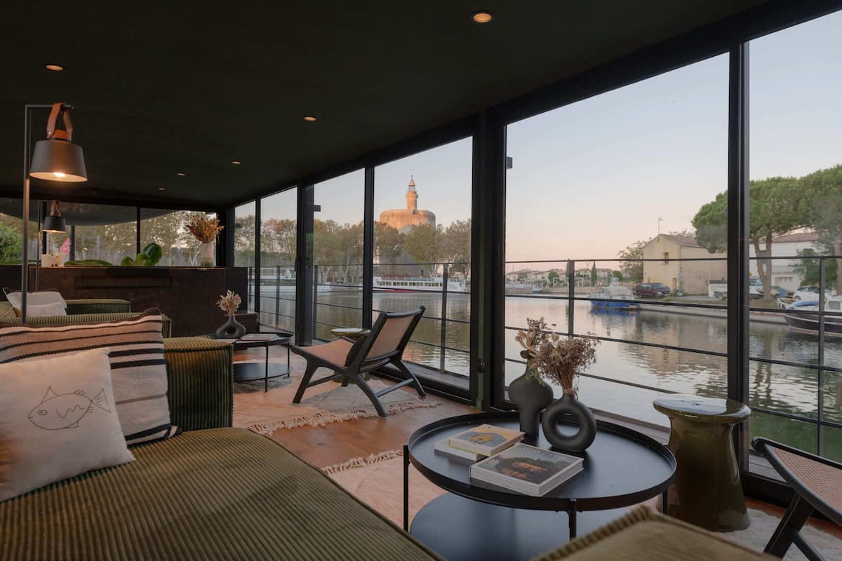 A modern interior with large windows overlooking a canal, featuring a sofa, chairs, a small table, and a bookshelf.