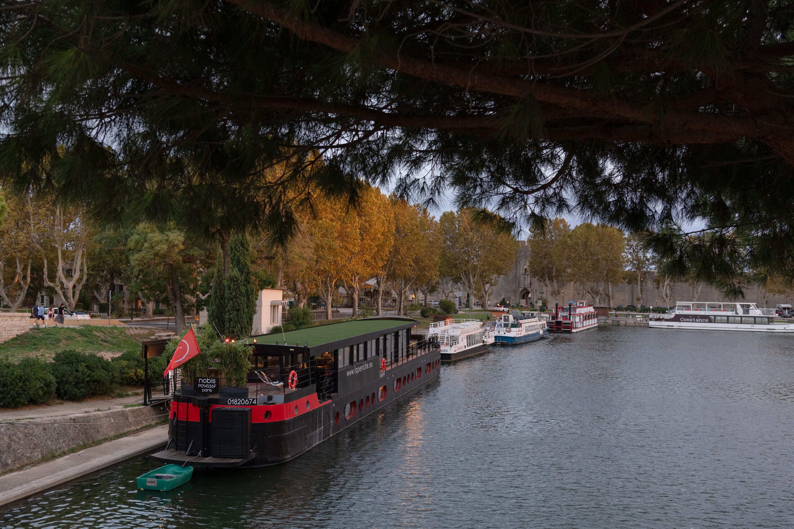 A river with boats, including a black boat with a red flag and a white boat with "Constance" written on it.