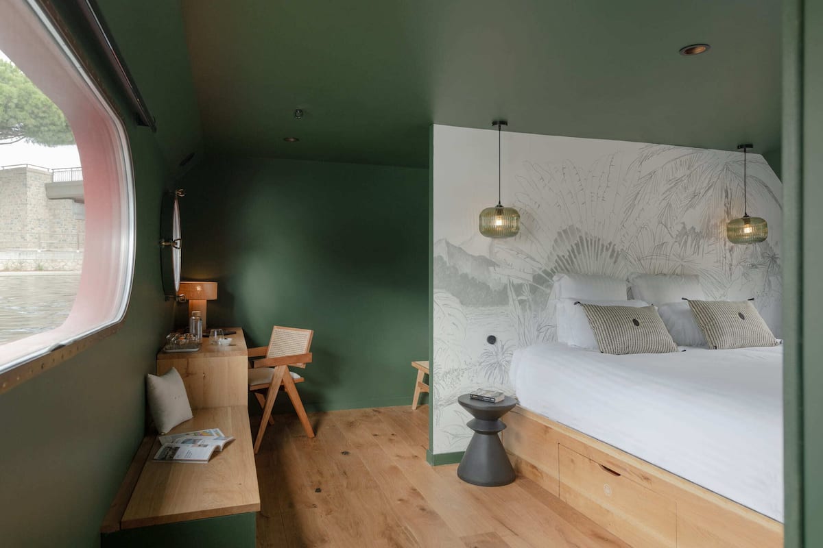 A bedroom with a bed, bedside table, lamp, and a view of the outside.