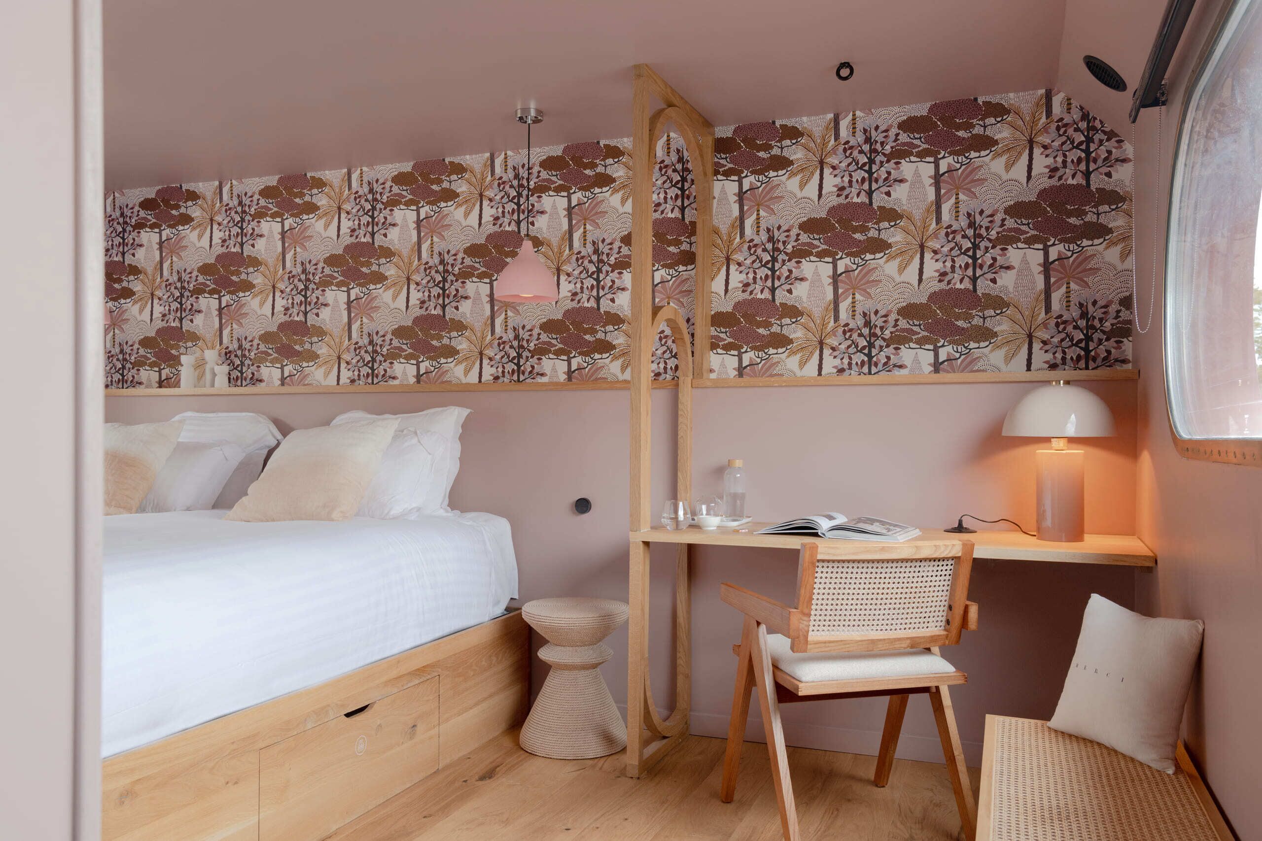 A bedroom with a bed, a desk, a chair, a bedside table, and a wall with a floral wallpaper.