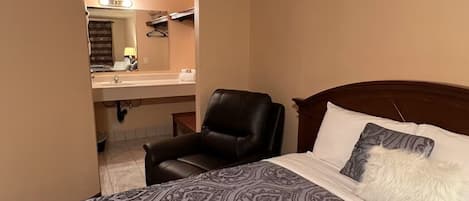 A hotel room with a bed, a chair, a vanity with a mirror, and a sink.