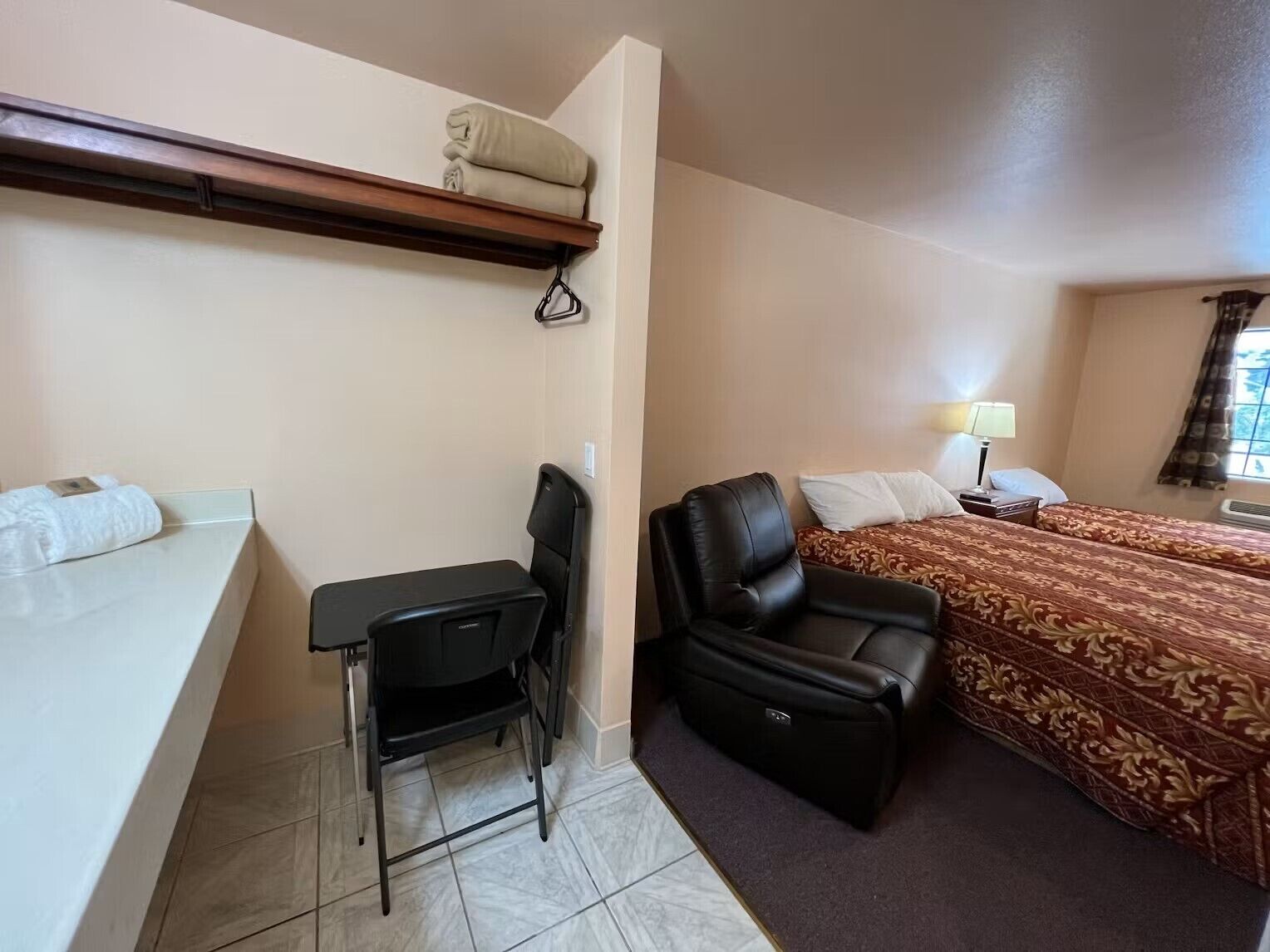 A hotel room with a bed, a desk with a chair, a small table, and a shelf with towels.