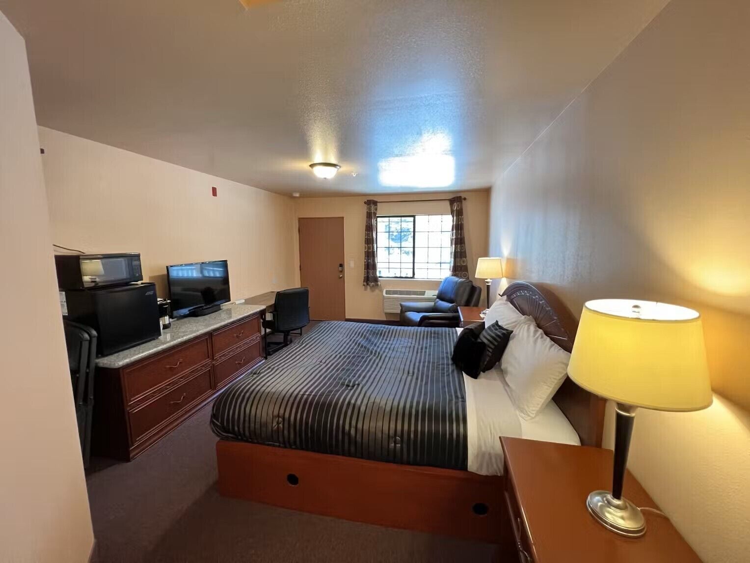 Habitación de hotel con cama, escritorio con computadora y televisor, una silla, lámpara y ventana con cortinas.