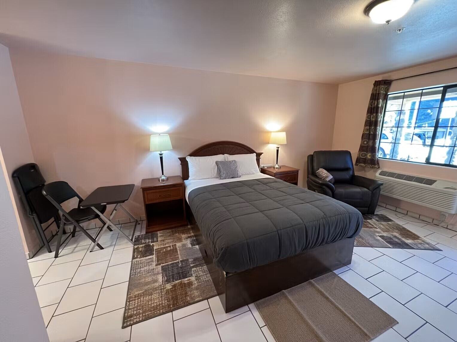 A hotel room with a bed, two bedside tables with lamps, a chair, a desk, and a window with a view of the street.
