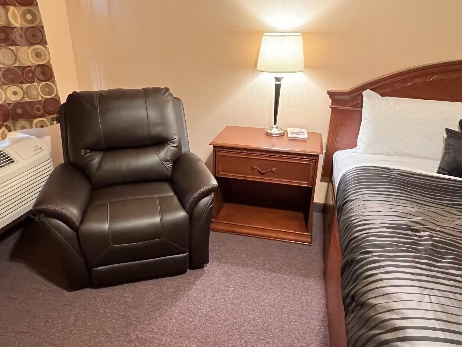 Habitación de hotel con un sillón de cuero, una mesita de noche con lámpara, una cama con ropa de cama a rayas y un papel tapiz de diseño circular en la pared.