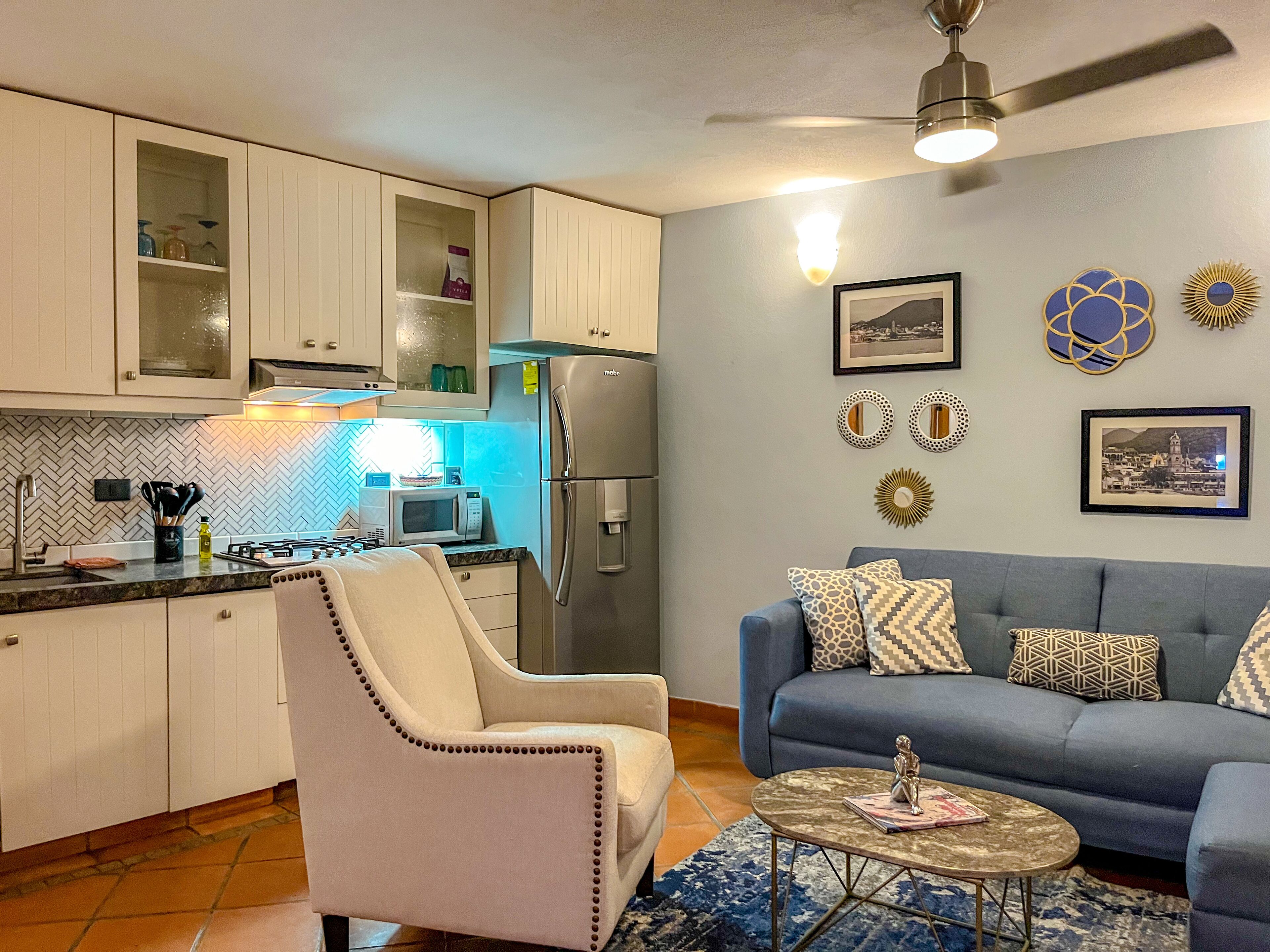 A compact kitchen with white cabinets, a microwave, and a refrigerator. There is a gray sofa with patterned cushions and a small round table with a book and a figurine.