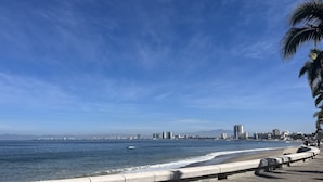 Un paisaje urbano costero con cielo despejado, una playa con barandilla de hormigón y un horizonte lejano.