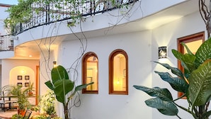 Un edificio blanco con ventanas arqueadas, un balcón con plantas y un patio con baldosas y mobiliario exterior.