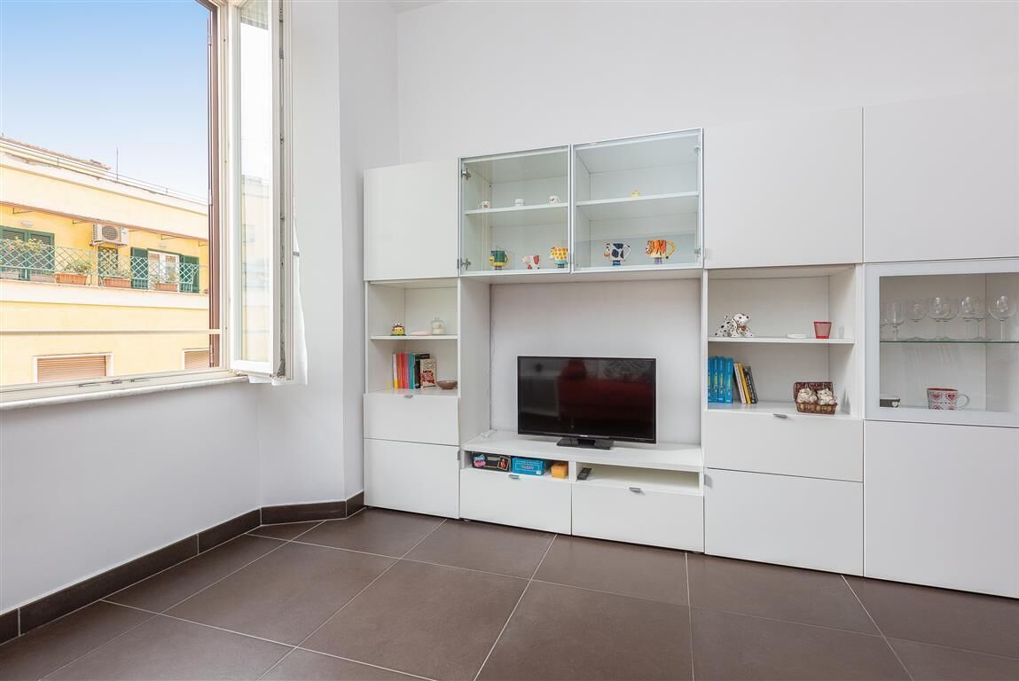 A modern living room with a flat-screen TV, built-in shelves, and a window overlooking a building.