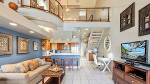 A spacious living area with a sofa, coffee table, and a TV. A kitchen is visible in the background, and there is a staircase leading to an upper level.