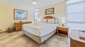 A hotel room with a large bed, a ceiling fan, a nightstand with a lamp, and artwork on the walls. - 1 bedroom Beachfront Condo, AC, Lanai Oceanview, 6 mile sandy beach (Kihei)
